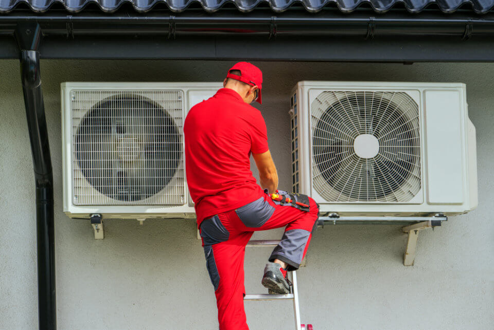 Contractor in Red Uniform on Ladder With Heat Pumps